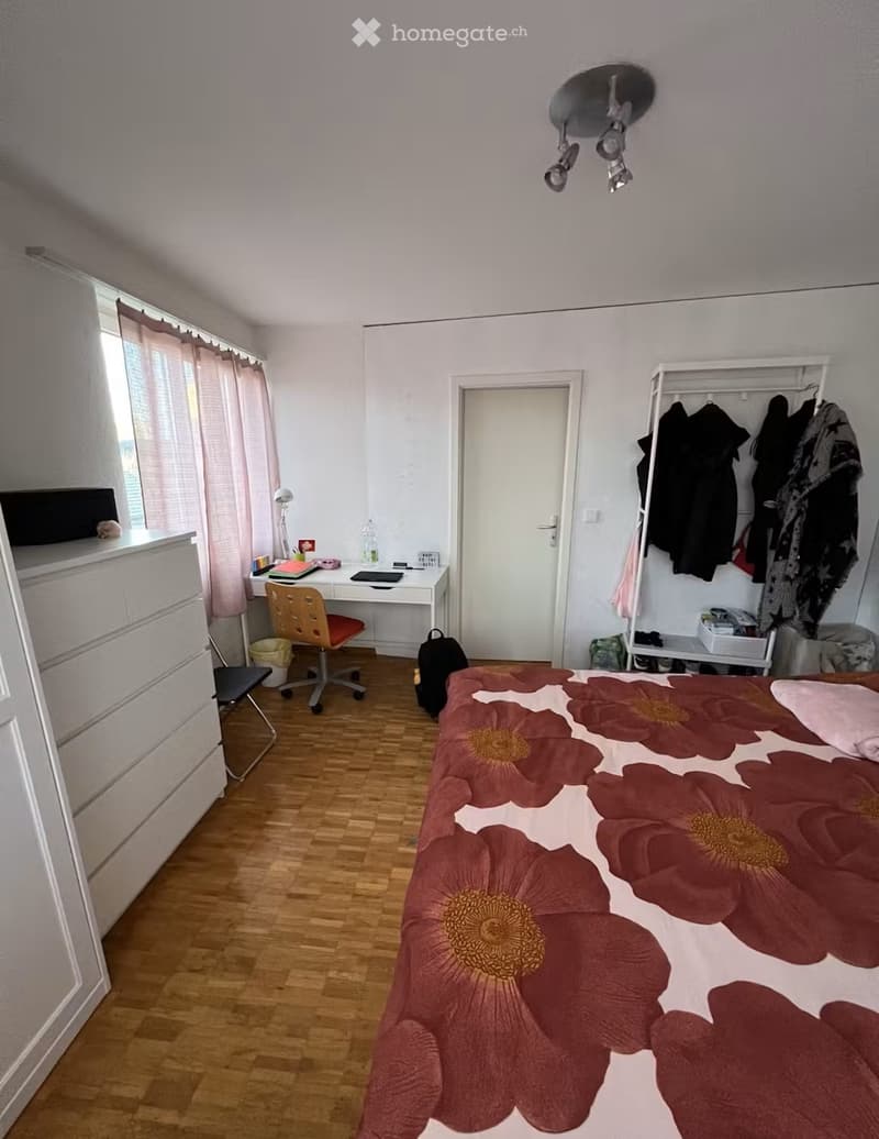 A well-lit bedroom with a wooden floor, a bed with a floral-patterned duvet, a desk with a chair, a wardrobe with hanging clothes, and a window with curtains.