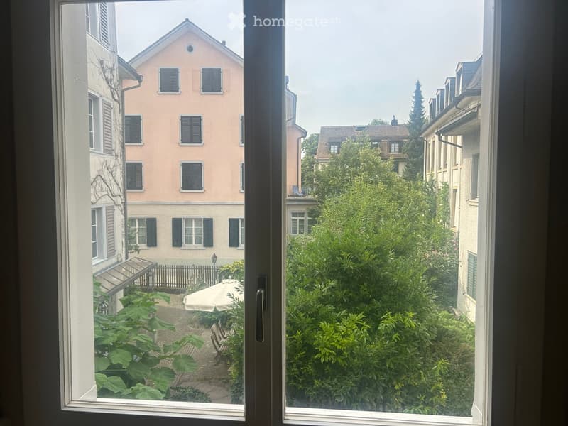View from a window showing a garden area with greenery, a small patio, and surrounding buildings.