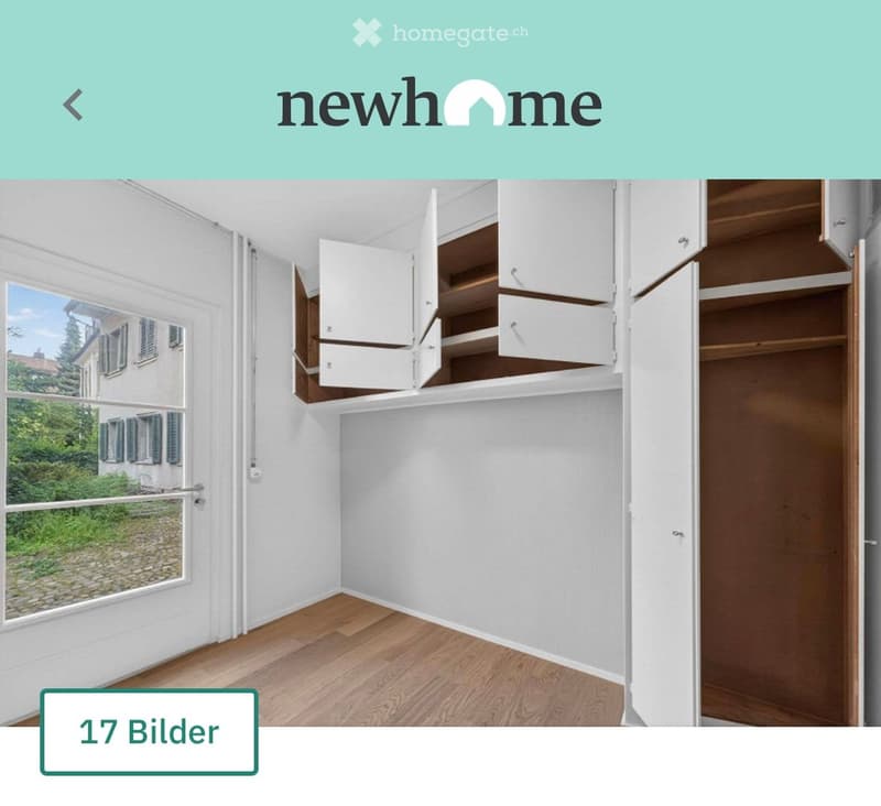Empty room with wooden flooring, white walls, large window with view of greenery, and built-in white cabinets with wooden shelves.