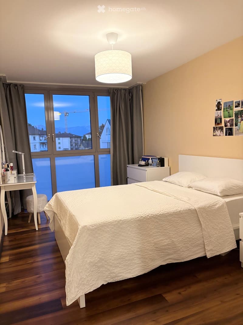 Modern bedroom with a large bed, nightstand, small table, and a view through large windows.