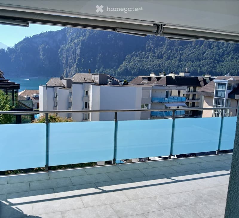 Balcony with a modern glass railing, overlooking a scenic lake and mountain landscape with surrounding buildings.