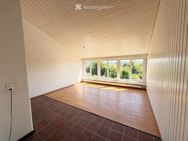 Empty room with wooden flooring and paneled walls, featuring large windows with a view of greenery outside.