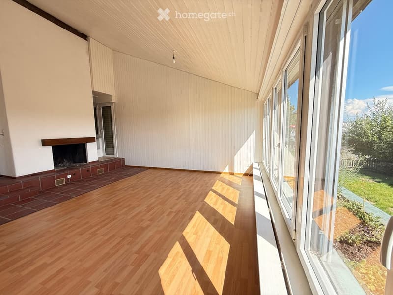Bright living room with wooden flooring, large windows offering a view, and a fireplace with a brick surround.