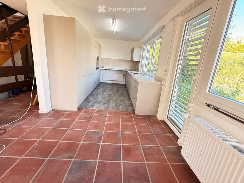 Empty kitchen with white cabinets, tiled floors, and large windows overlooking greenery. No furniture or appliances are present.