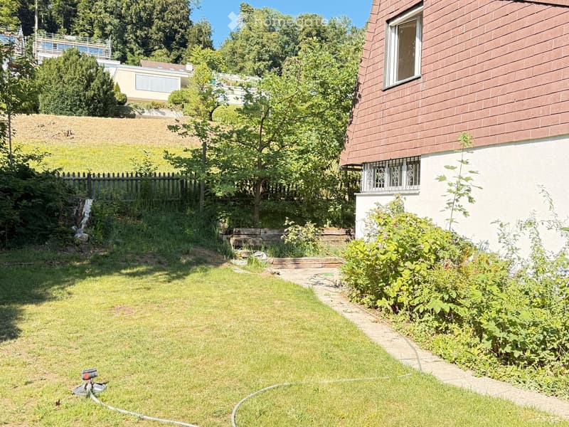 Well-maintained garden with a grassy lawn, a small waterfall, a wooden bench, and a modern house with a brown roof and white walls in the background.