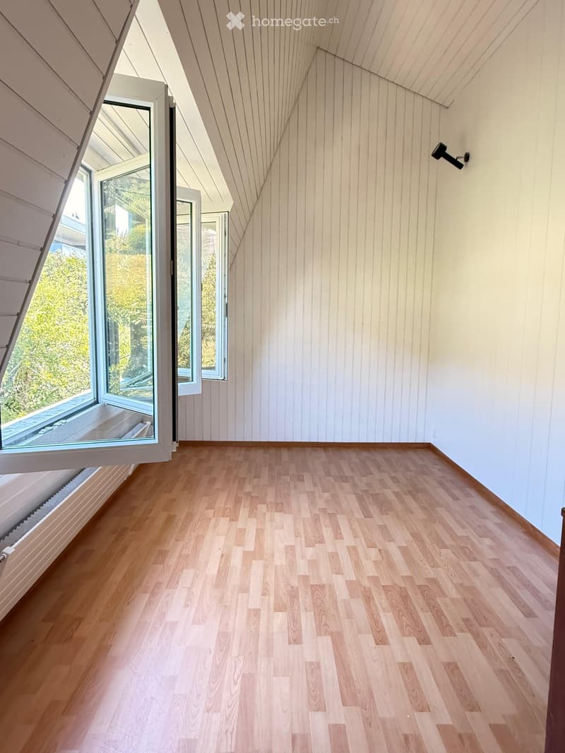 Empty room with wooden flooring, white paneled walls, large windows, and a slanted ceiling.