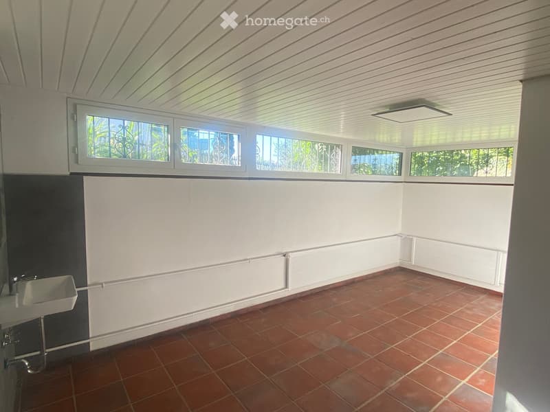 Empty room with red tiled floor, white walls, and a sink in the corner. Large windows with metal bars allow natural light to enter.