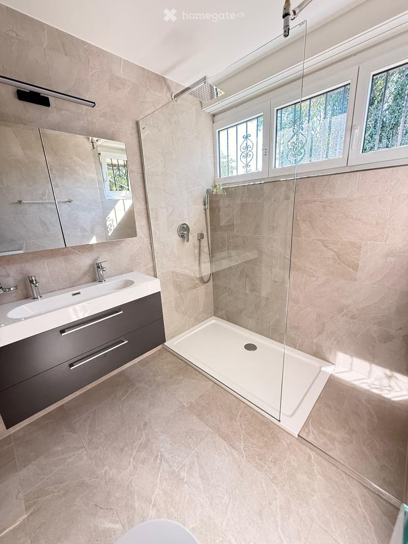 Modern bathroom with beige tiles, a large mirror, a white sink with a chrome faucet, and a walk-in shower with a glass enclosure and a handheld showerhead.