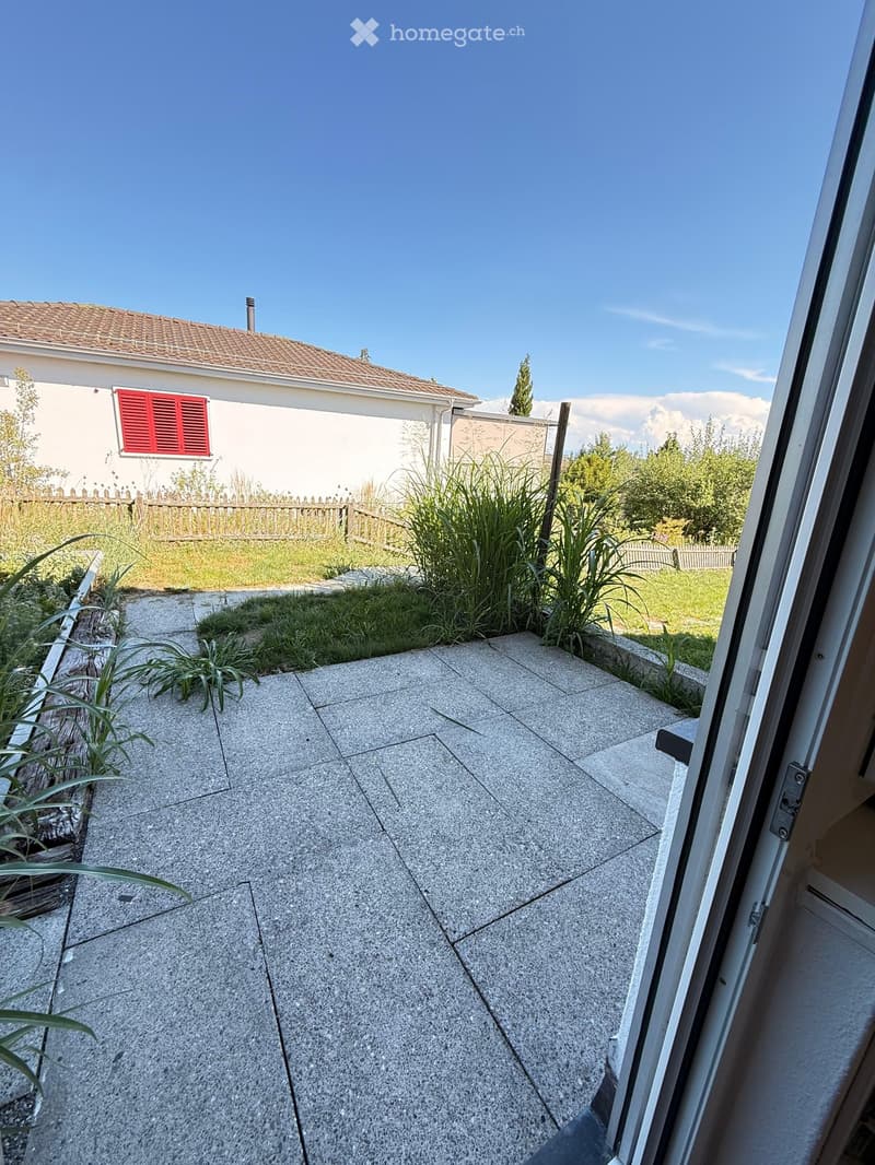 Balcony with concrete tiles, small garden area with plants and grass, view of a house with red shutters and a wooden fence.