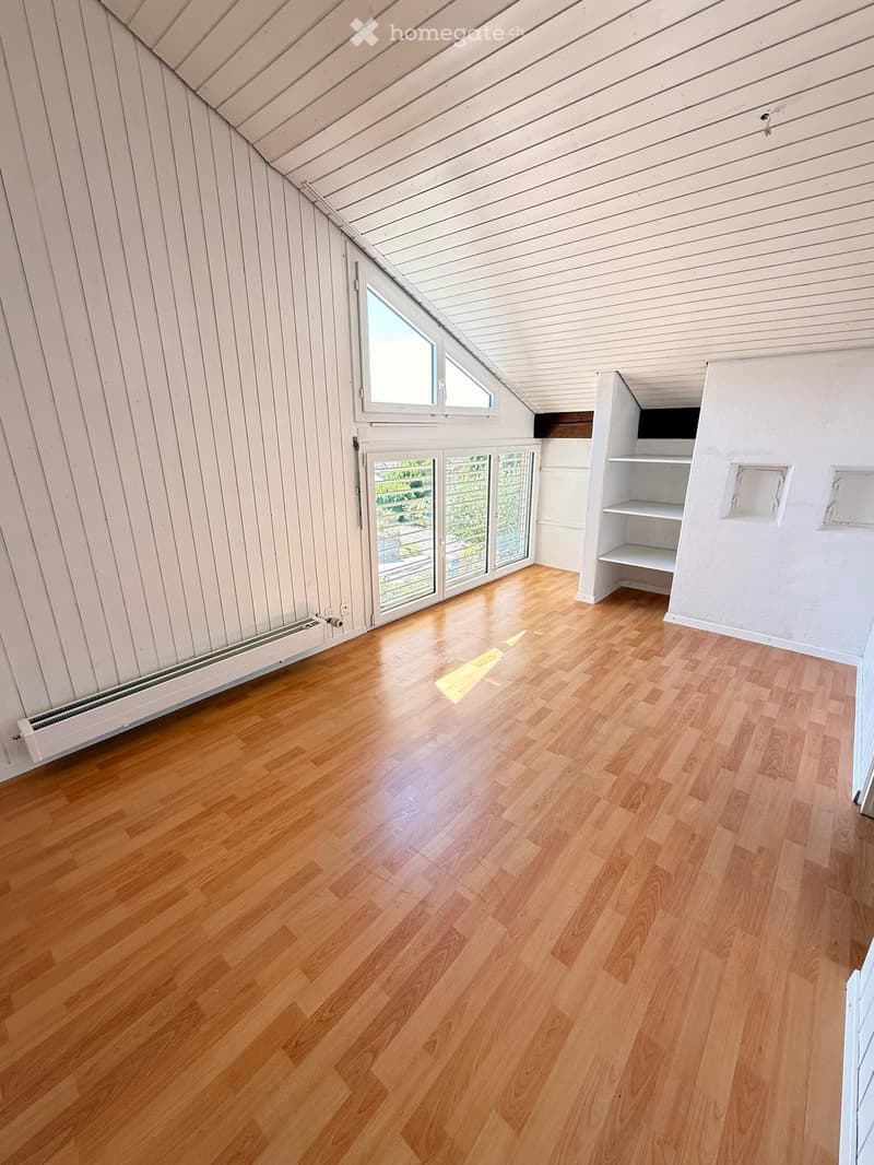 Empty attic room with wooden flooring, white paneled walls, and a skylight window. Features built-in shelving and a sliding glass door leading to a balcony.