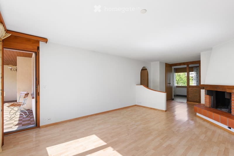 Bright, open living space with wooden flooring, white walls, and a fireplace. A sliding glass door leads to a patio area with outdoor furniture.