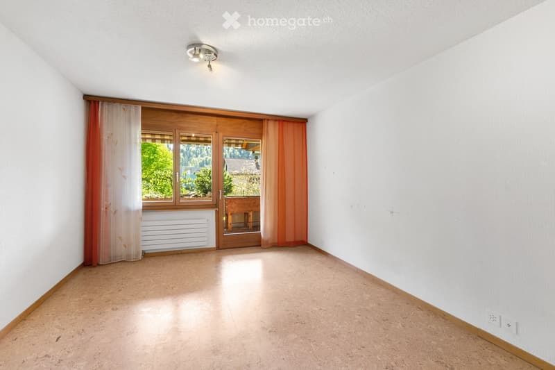 Empty room with light-colored walls, wooden flooring, and a large window with wooden frames and sheer curtains. The room has a ceiling light fixture and a radiator below the window.