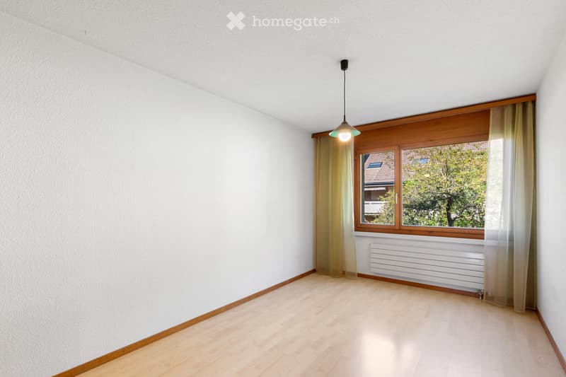 Empty room with light wooden flooring, white walls, large window with wooden frame and curtains, radiator below the window, and a hanging light fixture.