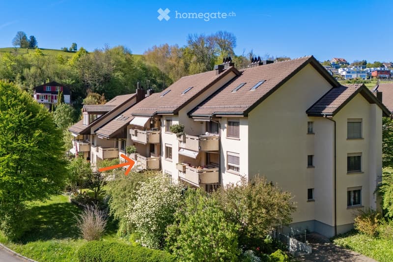 A multi-story residential building with a brown tiled roof, multiple balconies, and surrounding greenery. The building is situated in a hilly, landscaped area with trees and shrubs.