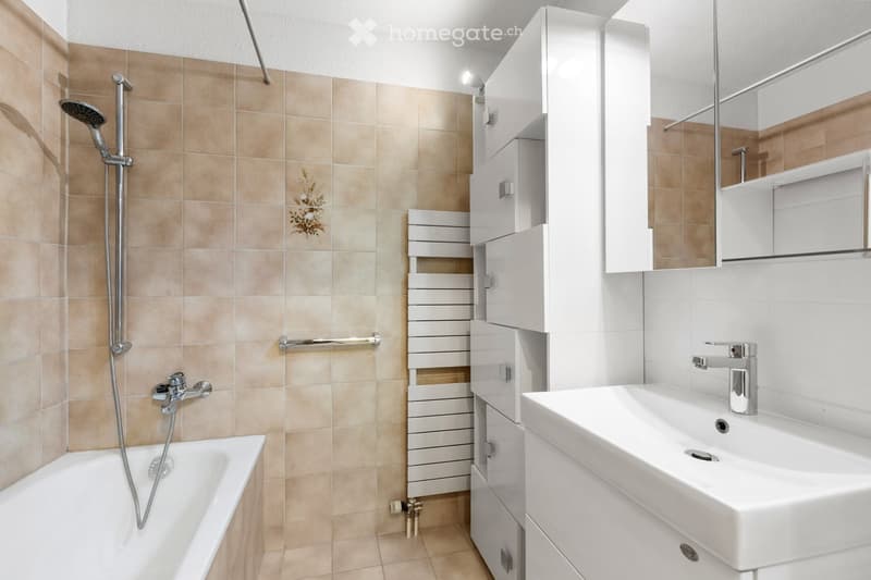 Modern bathroom with beige tiled walls, white bathtub, sink, mirror, and built-in cabinets.
