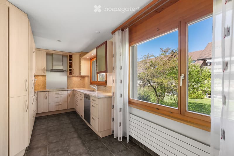 Modern kitchen with light wood cabinets, stainless steel appliances, granite countertops, and large windows overlooking a green area.
