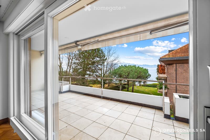 Spacious balcony with tiled flooring, white railings, and a scenic view of a green lawn, trees, and a body of water under a clear blue sky.