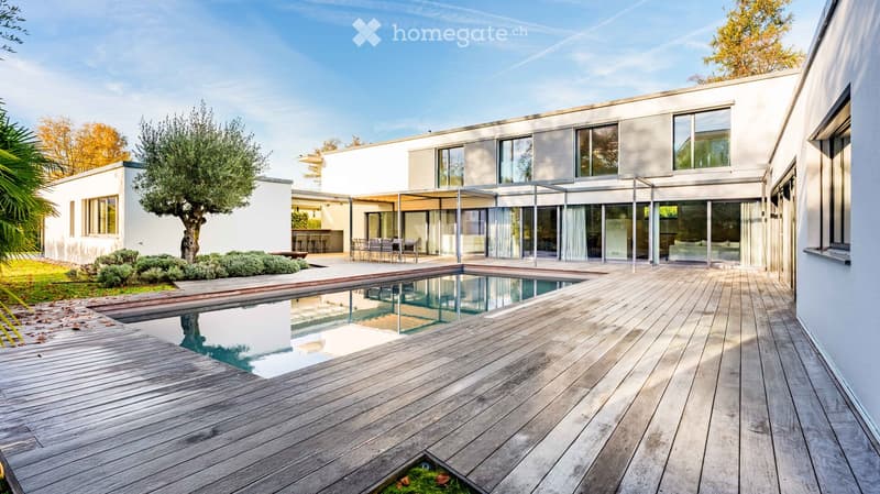 Modern house with a swimming pool, wooden decking, outdoor seating area, and surrounding greenery.