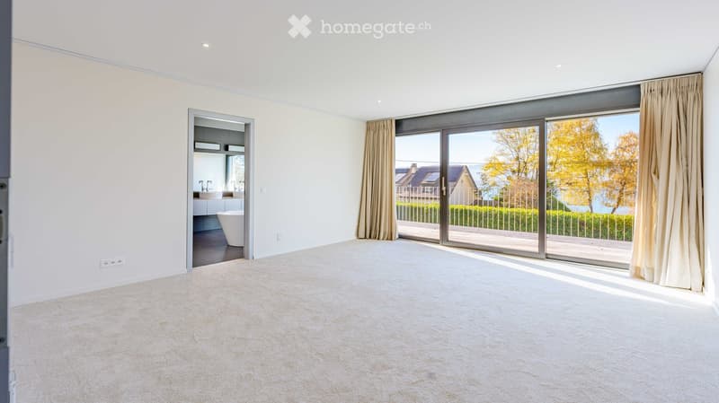 Empty room with white walls, carpeted floor, large sliding glass doors leading to a balcony with a scenic view of the lake and surrounding houses.