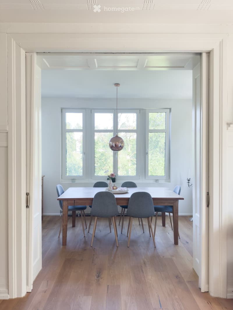 Bright dining room with wooden floor, large windows, modern dining table with six chairs, vase with flowers, and candle holder.