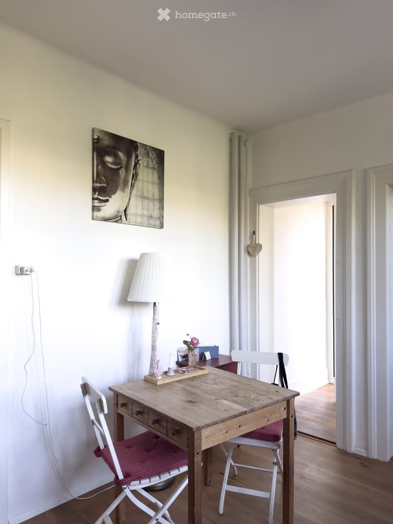A small, well-lit room with a wooden desk, two chairs, a lamp, and a picture on the wall.
