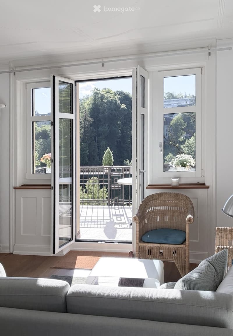 Bright living room with large windows and sliding doors opening to a balcony, featuring a grey sofa, white coffee table, and wicker chair.