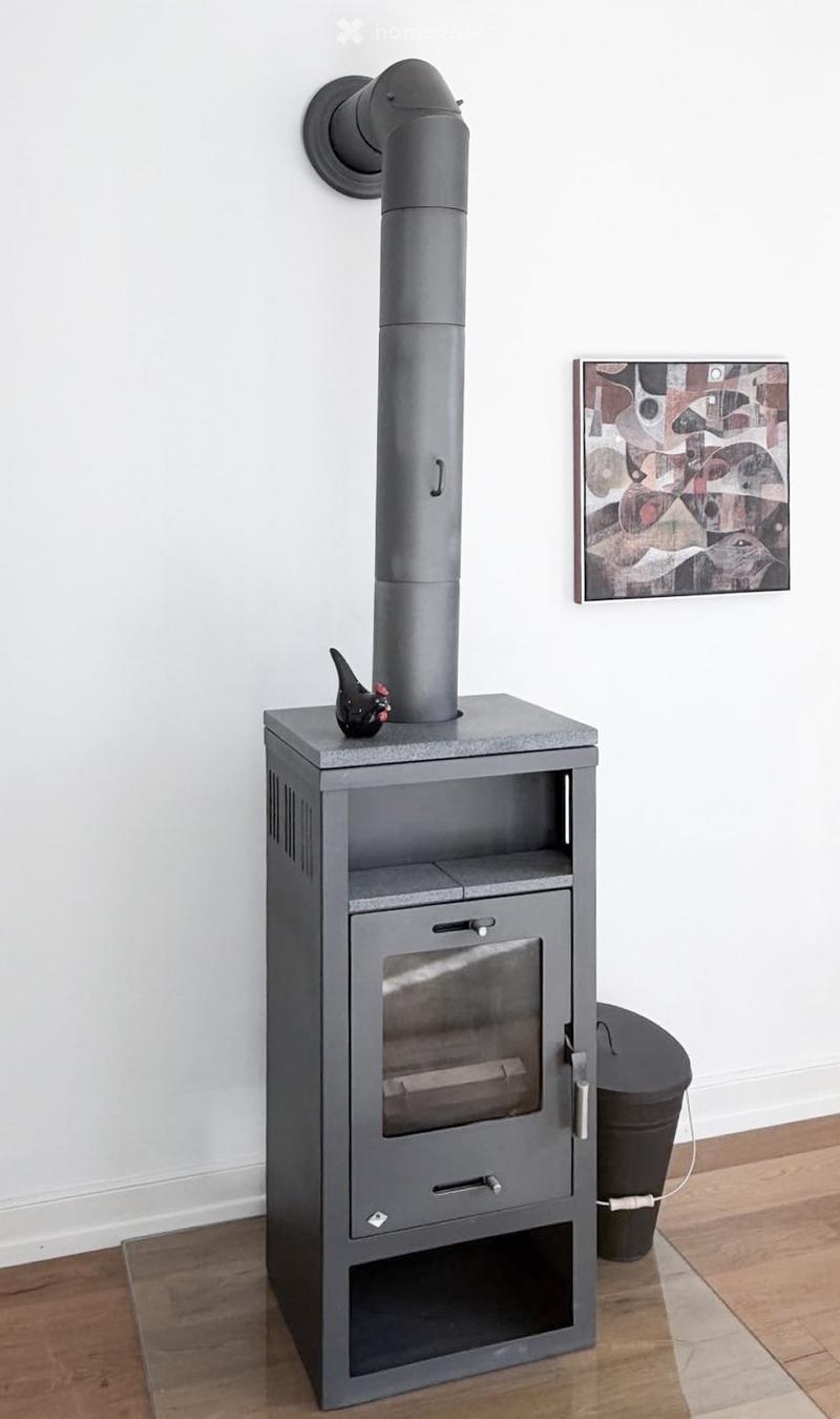 A modern wood-burning stove with a tall chimney, a decorative bird figurine on top, a picture frame on the wall, and a trash can nearby.