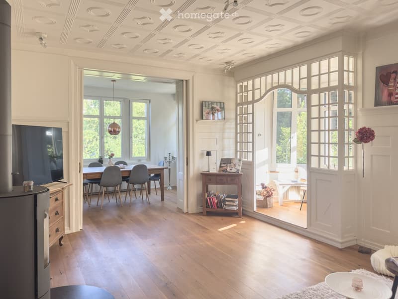 Open-plan living and dining area with wooden flooring, large windows, a fireplace, a TV, a dining table with chairs, a bookshelf, and decorative items.