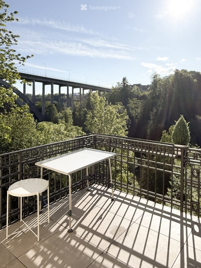 Balcony with a metal railing, a white table, and a stool, overlooking a scenic view of a bridge and lush greenery.
