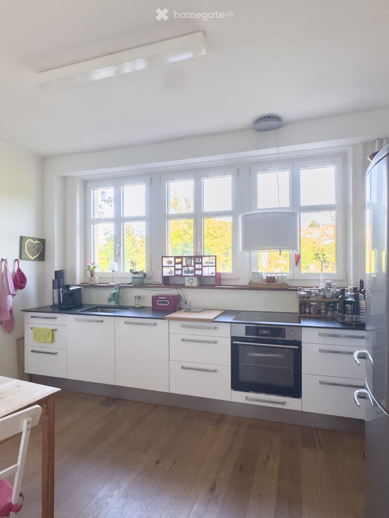 Bright, modern kitchen with white cabinets, wooden flooring, and large windows. Features include a refrigerator, oven, sink, coffee maker, and various kitchen utensils.