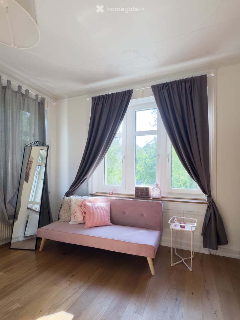 A cozy, well-lit room with wooden flooring, a pink upholstered sofa with decorative pillows, a small white side table with a cup, a mirror, and dark curtains covering the windows.