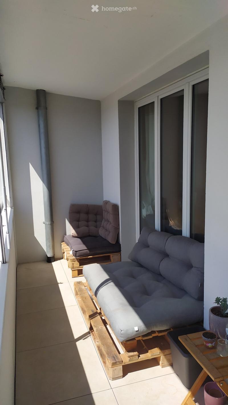 Balcony with a grey sofa, wooden chair, small table, and potted plants.