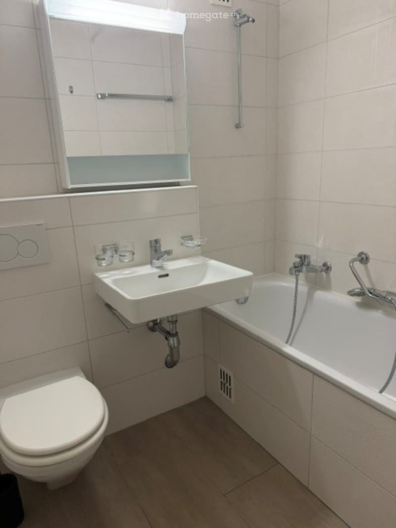 Modern bathroom with white tiles, a corner sink, a bathtub, a toilet, and a mirrored cabinet above the sink.