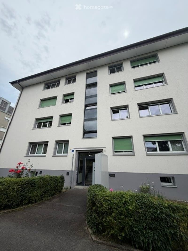 Modern multi-story residential building with white facade, green window frames, and neatly trimmed hedges in front.