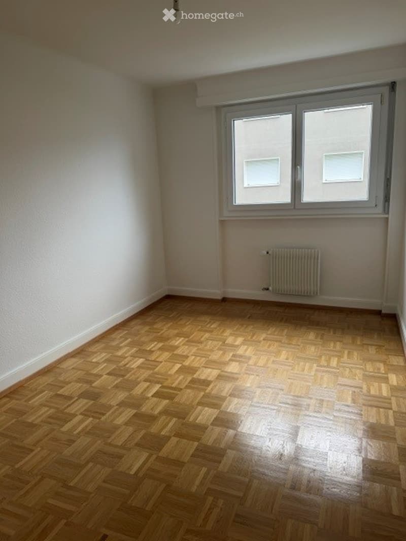 Empty room with wooden parquet flooring, white walls, and a single window with white frame and radiator below.