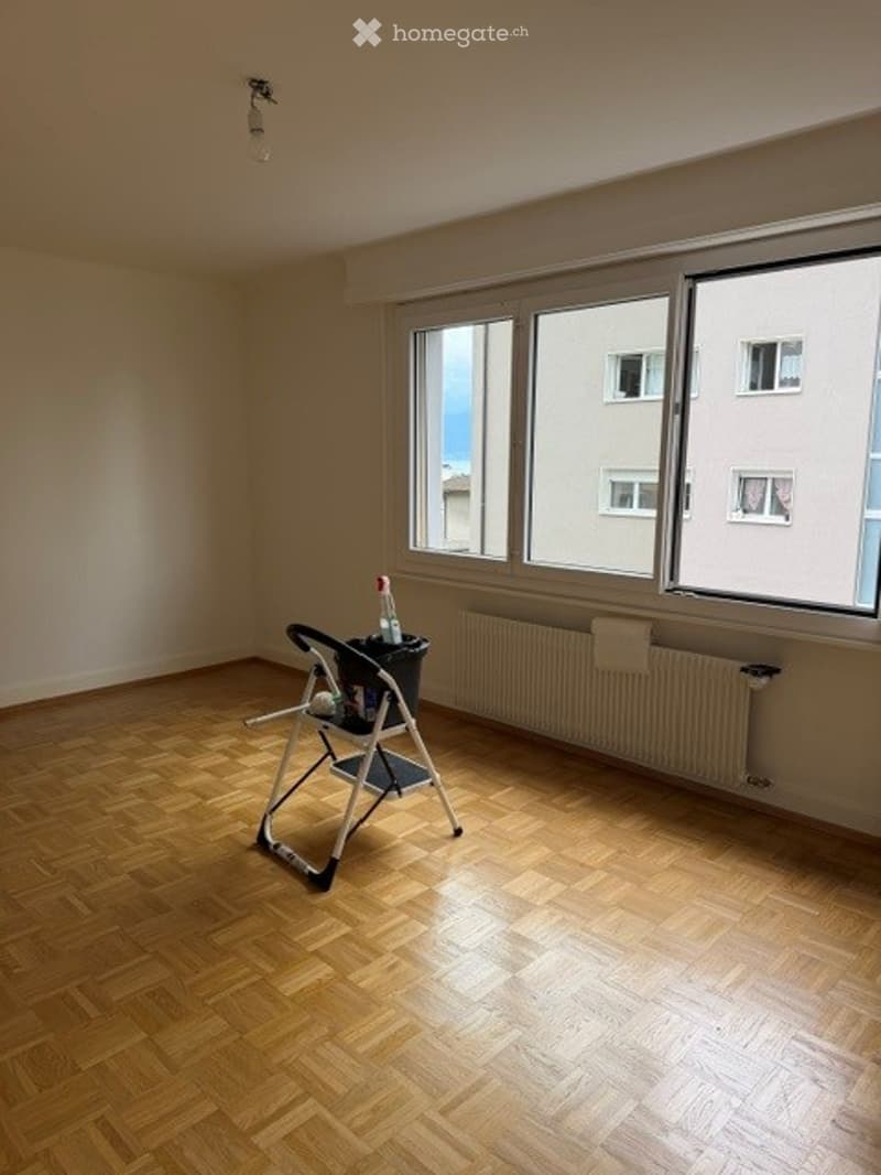 Empty room with wooden flooring, large windows, radiator, and a small folding chair with a bucket on it.