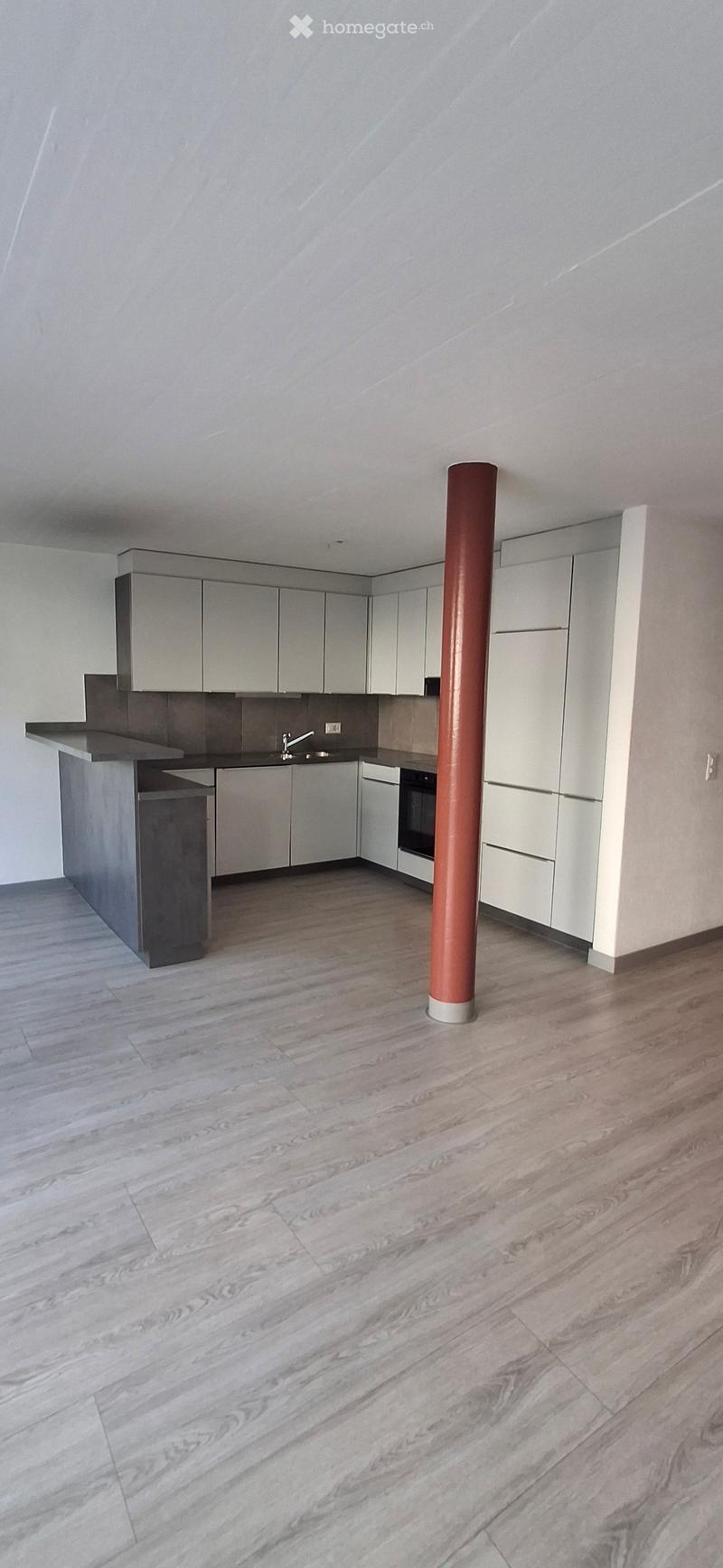 Modern kitchen with light grey cabinets, a central island, and a sink with a faucet. The floor is made of light wood, and there is a red column in the center.