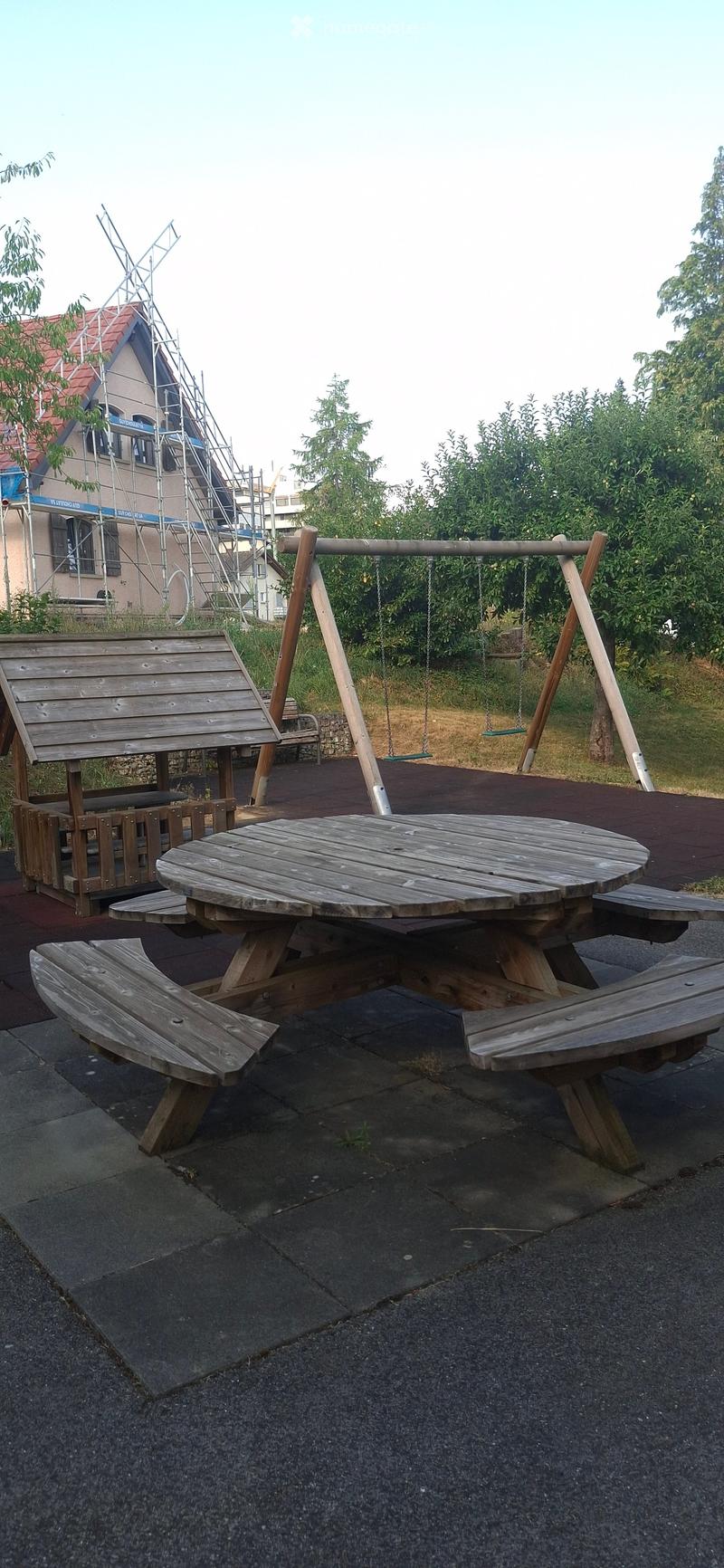 Outdoor area with a wooden picnic table, benches, and a swing set in the background.