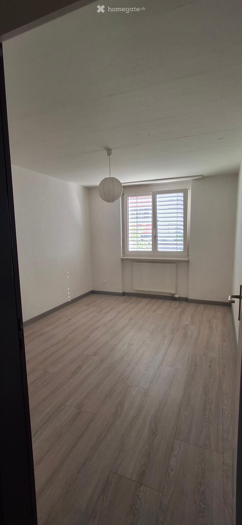 Empty room with wooden flooring, white walls, a single window with blinds, a radiator, and a hanging light fixture.