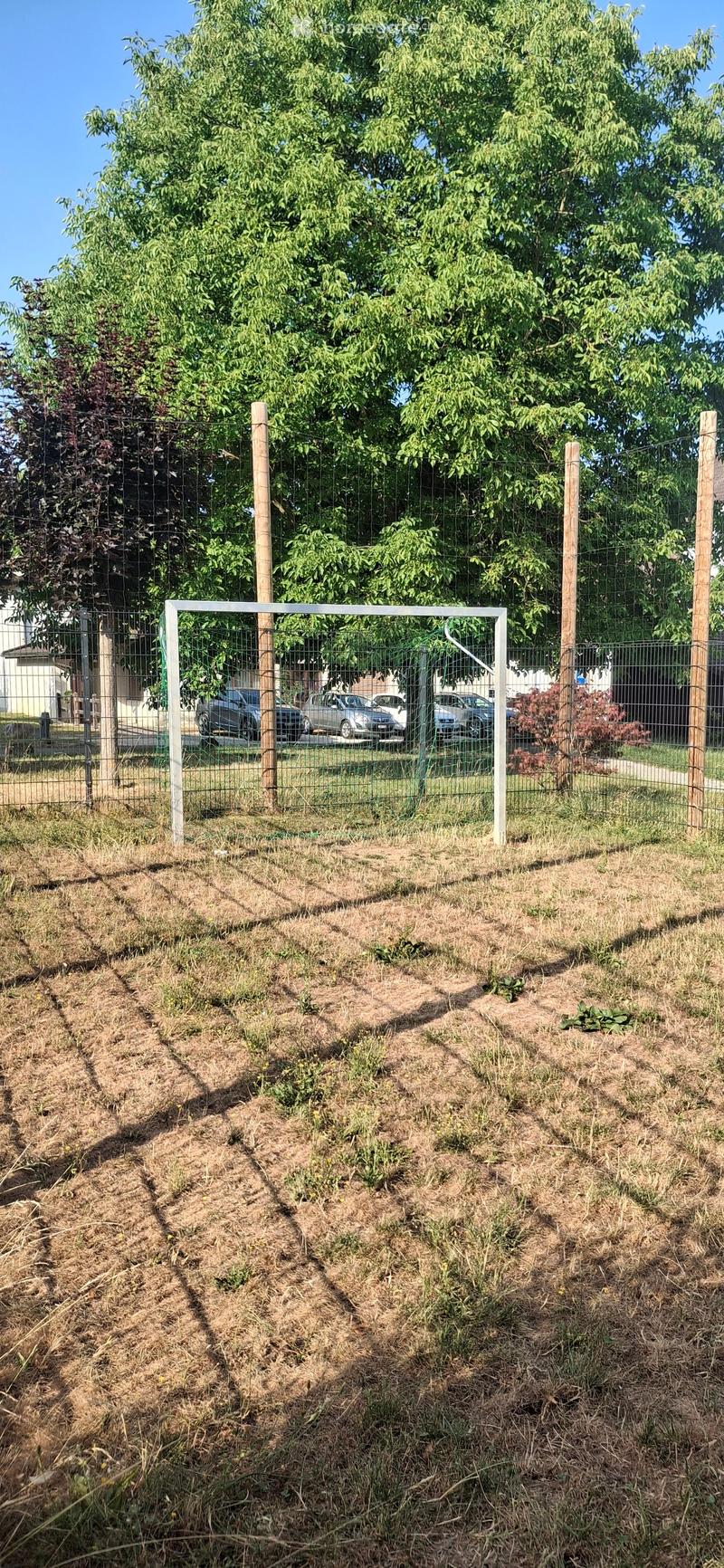 A fenced garden area with raised beds covered in straw, surrounded by trees and parked cars in the background.
