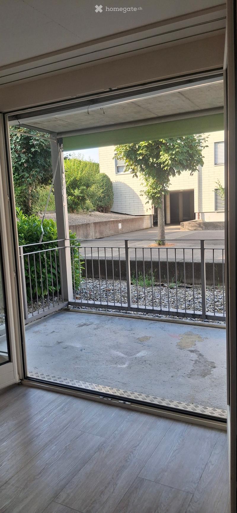 Open balcony with wooden flooring, metal railing, and a view of a garden area with trees and a building in the background.