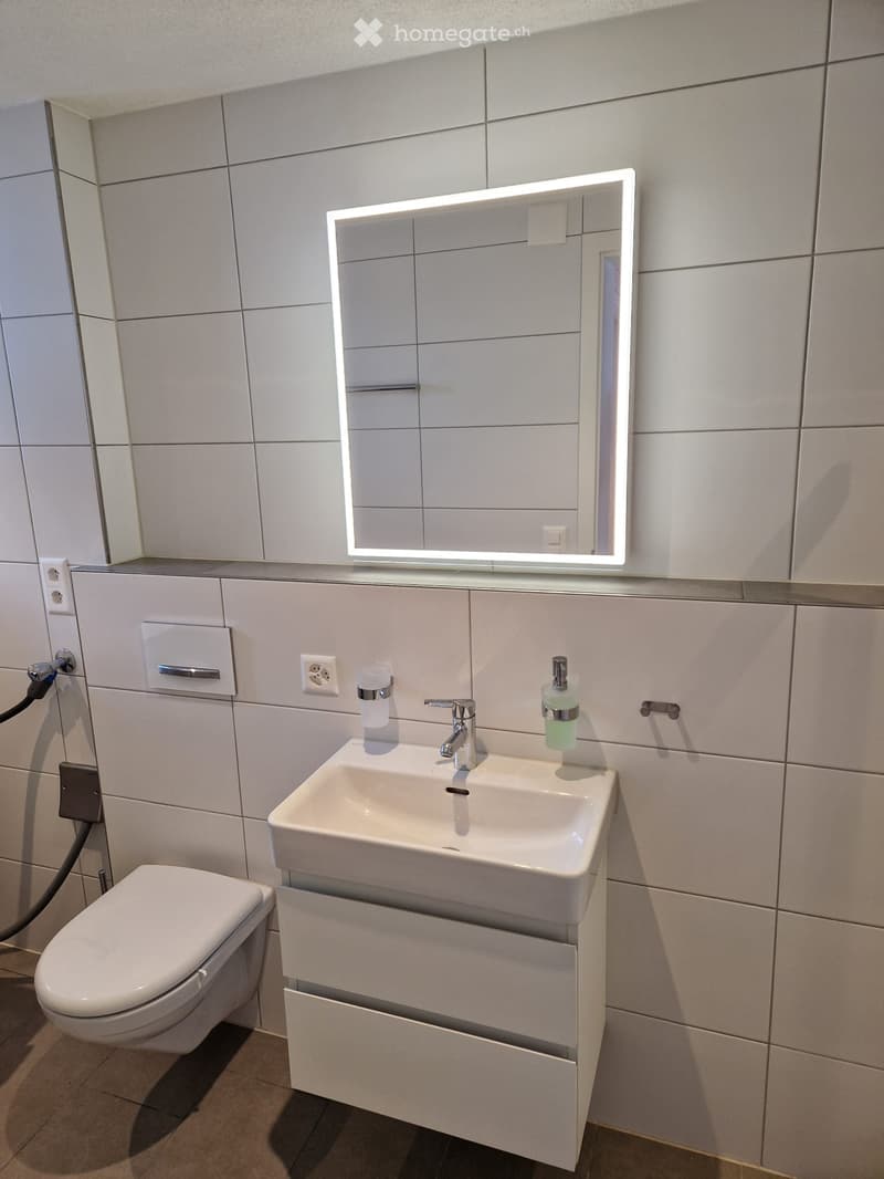 Modern bathroom with white tiles, a white sink, a mirror with a light, a toilet, and a power outlet