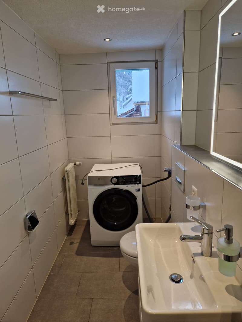 tiled walls and floor, white sink, white washing machine, window, toilet, white mirror