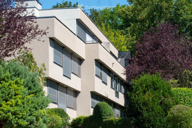 Modern multi-story building with large windows and shutters, surrounded by lush greenery and trees.