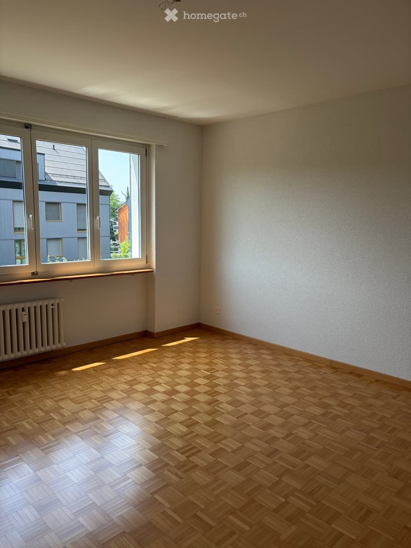Empty room with wooden herringbone flooring, white walls, large windows, and a radiator under the windows.