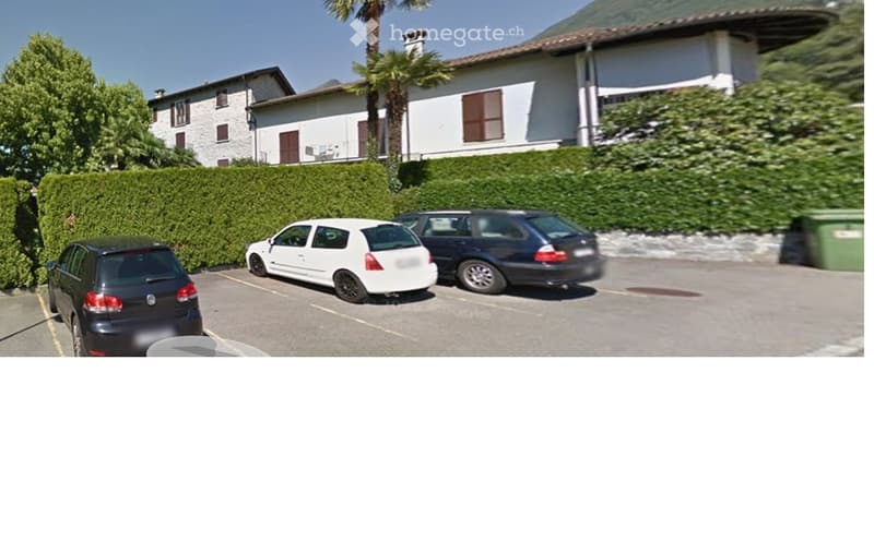 A residential building with a parking area in front, featuring several parked cars and a well-maintained hedge lining the driveway.
