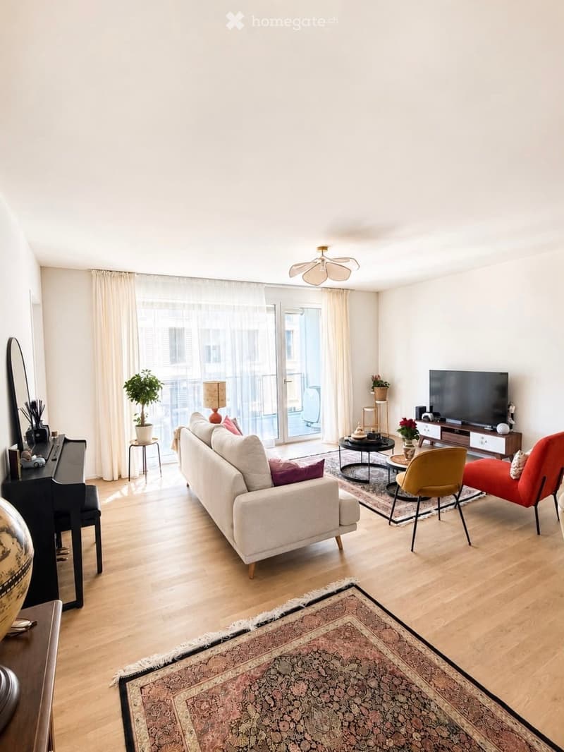 Modern living room with light wooden flooring, large windows with white curtains, a white sofa with colorful cushions, a coffee table, a red armchair, a yellow chair, a piano, a television, and decorative plants.
