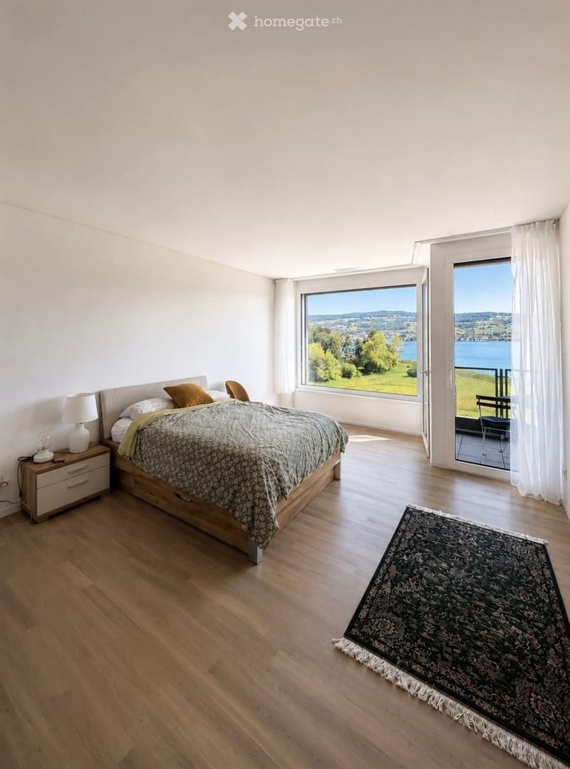 Bright bedroom with wooden flooring, large windows offering scenic views, a bed with patterned bedding, a nightstand with a lamp, and a decorative rug.