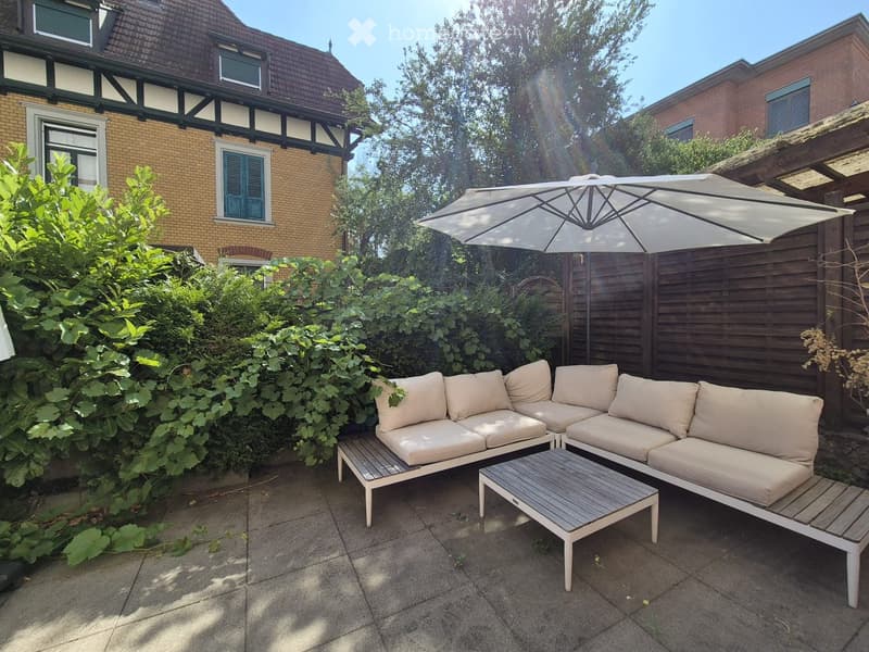 Garden area with a patio, beige outdoor sofa set, coffee table, umbrella, and surrounding greenery.