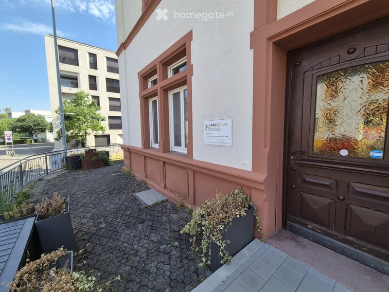 Entrance to a building with a cobblestone pathway, potted plants, and a sign for 'PROPERIUM' property management.